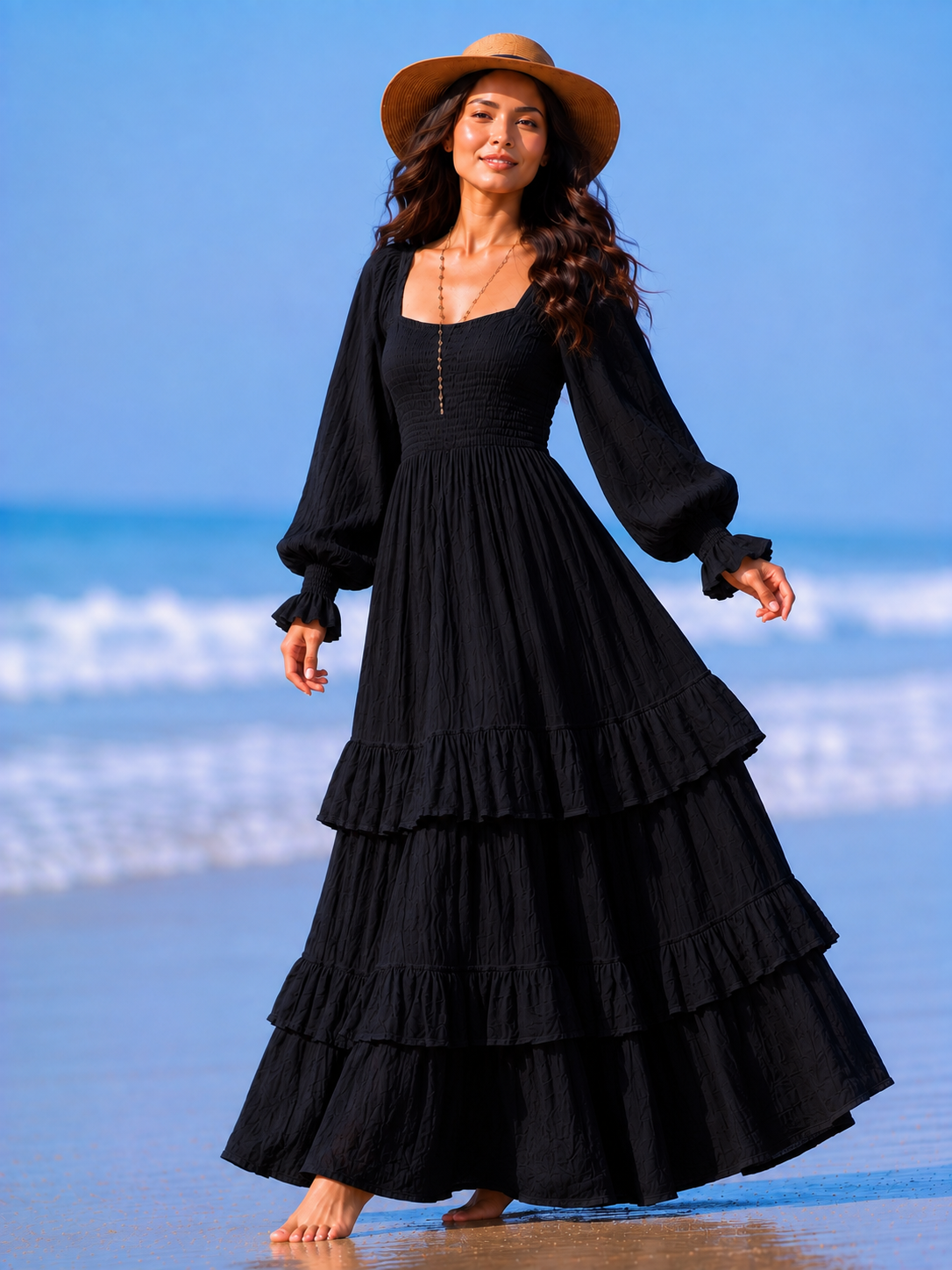 A woman stands barefoot on a beach wearing a long black tiered maxi dress with long balloon sleeves and a wide-brim tan hat.