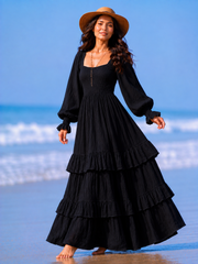 A woman stands barefoot on a beach wearing a long black tiered maxi dress with long balloon sleeves and a wide-brim tan hat.